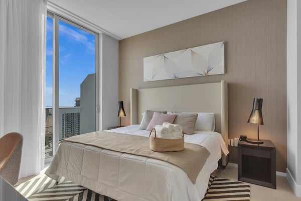 Modern bedroom with upholstered headboard, floor-to-ceiling windows overlooking city skyline, and neutral beige and white furnishings.