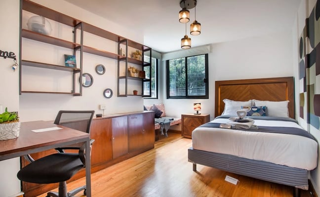Modern bedroom with queen bed, wood desk, open shelving, pendant lights, and forest views through large windows